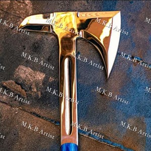 May include: A polished, reflective metal axe head with a sharp, curved blade and a pointed rear spike. The surface is highly reflective, showing golden highlights. The text M.K.B Artist is repeated across the image. A blue handle section is visible.