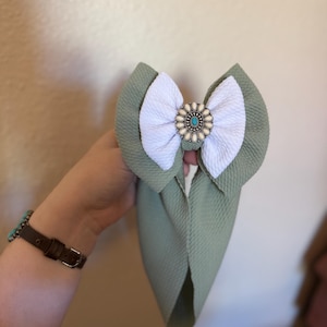 May include: A sage green and white baby bow with a turquoise and cream-colored center embellishment. The bow is made of textured fabric and is designed to be worn on the head. The bow is a stylish accessory for babies and young children.