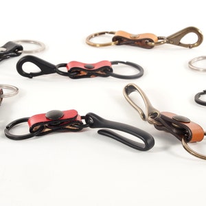 May include: A collection of six leather keychains with different colored leather straps and metal rings. The keychains feature a variety of metal hooks and clasps.