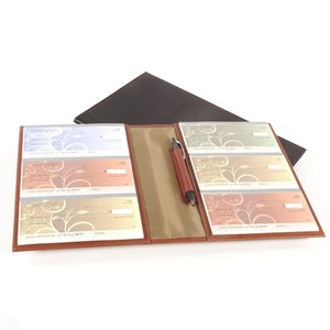 May include: A brown leather checkbook holder with six checkbook slots and a pen holder. The holder is open and the checkbooks are visible. The checkbooks have a floral design.