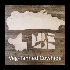 May include: A large, irregularly shaped piece of light tan veg-tanned cowhide, and several smaller cut pieces, are displayed on a wooden surface. The text "Veg-Tanned Cowhide" is printed in white at the bottom of the image.