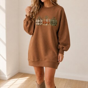 May include: A brown long-sleeve sweatshirt dress featuring a crew neckline and ribbed cuffs. The front of the garment displays a graphic print of three pumpkins, each decorated with a distinct plaid pattern in shades of brown, orange, and green.