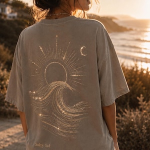 Op de afbeelding: Een taupekleurig t-shirt met korte mouwen en een gouden afbeelding op de achterkant. Het ontwerp toont een gestileerde zon die opkomt boven een grote oceaangolf met een maansikkel aan de rechterkant. De tekst Salty Sol staat onderaan in kleine gouden letters.