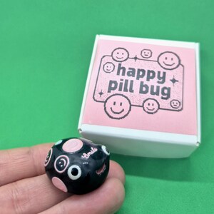 May include: A black and pink pill bug with white eyes and a smiling face. The pill bug is next to a white box with the words "happy pill bug" and several smiley faces printed on it. The background is a solid green colour.