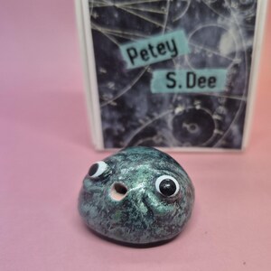 May include: A small, rounded, metallic teal-coloured ceramic sculpture with a textured finish, featuring two large googly eyes and a small circular opening. Behind it stands a rectangular card displaying the text Petey S. Dee on a dark background.