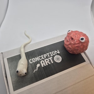 Peut inclure: Une sculpture blanche en forme de spermatozoïde et une sculpture sphérique rouge et texturée avec des yeux globuleux reposent sur une boîte. La boîte porte l'inscription "CONCEPTION ART". La sculpture de spermatozoïde a une queue incurvée.
