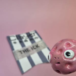 Puede incluir: Una escultura rosa texturizada con forma de alienígena con un ojo grande y un pequeño letrero borroso que dice "THE ICK". El letrero tiene rayas azules y blancas. El fondo es de color rosa suave.