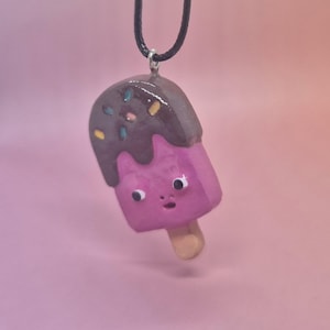 May include: A pendant shaped like a pink popsicle with a brown chocolate-dipped top featuring colorful sprinkles. The popsicle has a small face with two white eyes and a tiny mouth. It hangs from a black cord and has a light tan wooden stick base.