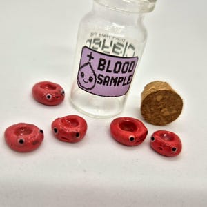 May include: A clear glass vial with a cork stopper and a label that reads "BLOOD SAMPLE." The label features a cartoon blood drop. Several small, red, cartoon blood cells with black eyes are scattered around the vial.