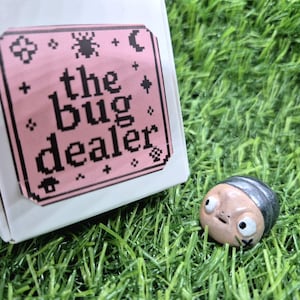 Puede incluir: Una pequeña figura de insecto de cerámica pintada a mano con una expresión de sorpresa, sobre hierba verde. Junto a ella, una caja blanca con una etiqueta rosa que dice "the bug dealer" en fuente negra pixelada.