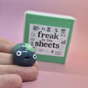 May include: A small, dark blue, rounded ceramic figure with two white eyes and a small mouth, positioned in front of a square green box. The box features a white label with black text that reads "freak in the sheets" and various spreadsheet icons.