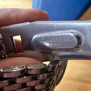 May include: Close-up of a stainless steel watch band clasp. The clasp is silver and has the words "STAINLESS STEEL CHINA" printed on it. The watch band is made of woven metal links.