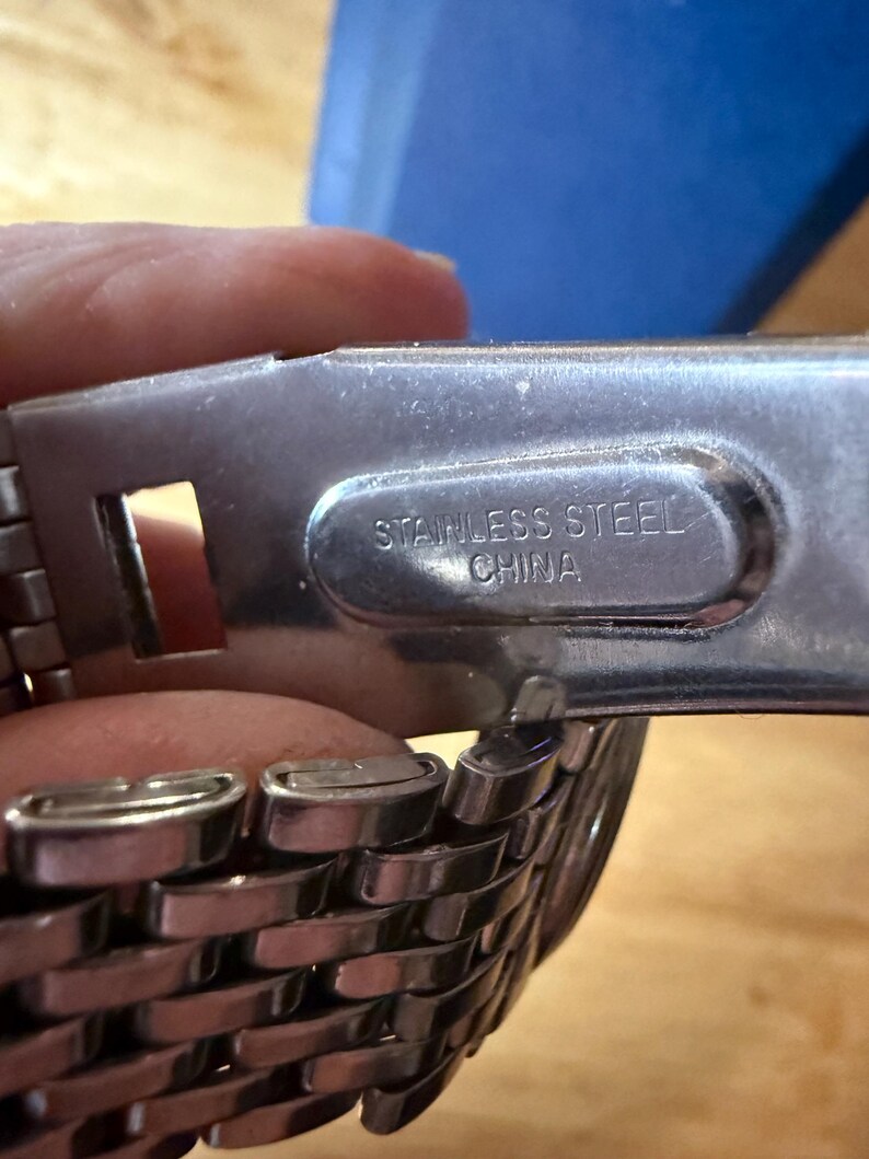 May include: Close-up of a stainless steel watch band clasp. The clasp is silver and has the words "STAINLESS STEEL CHINA" printed on it. The watch band is made of woven metal links.