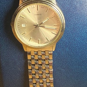 May include: A gold-toned quartz watch with a silver and gold band. The watch face is gold with black hands and markers. The watch displays the date and the word "QUARTZ".