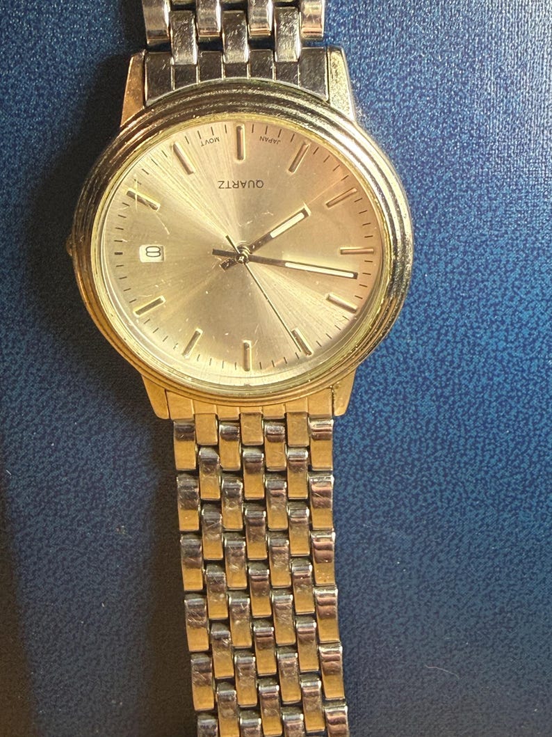 May include: A gold-toned quartz watch with a silver and gold band. The watch face is gold with black hands and markers. The watch displays the date and the word "QUARTZ".