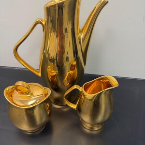 May include: A gold-colored tea set, including a teapot, a creamer, and a sugar bowl with a lid. The set has a shiny, reflective surface and a modern design. The teapot has a long spout and a curved handle.