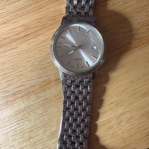 May include: Silver-toned quartz watch with a rectangular link band. The watch face is silver with black hour markers and hands. The word "QUARTZ" is printed on the watch face. The date is displayed at the 3 o'clock position.