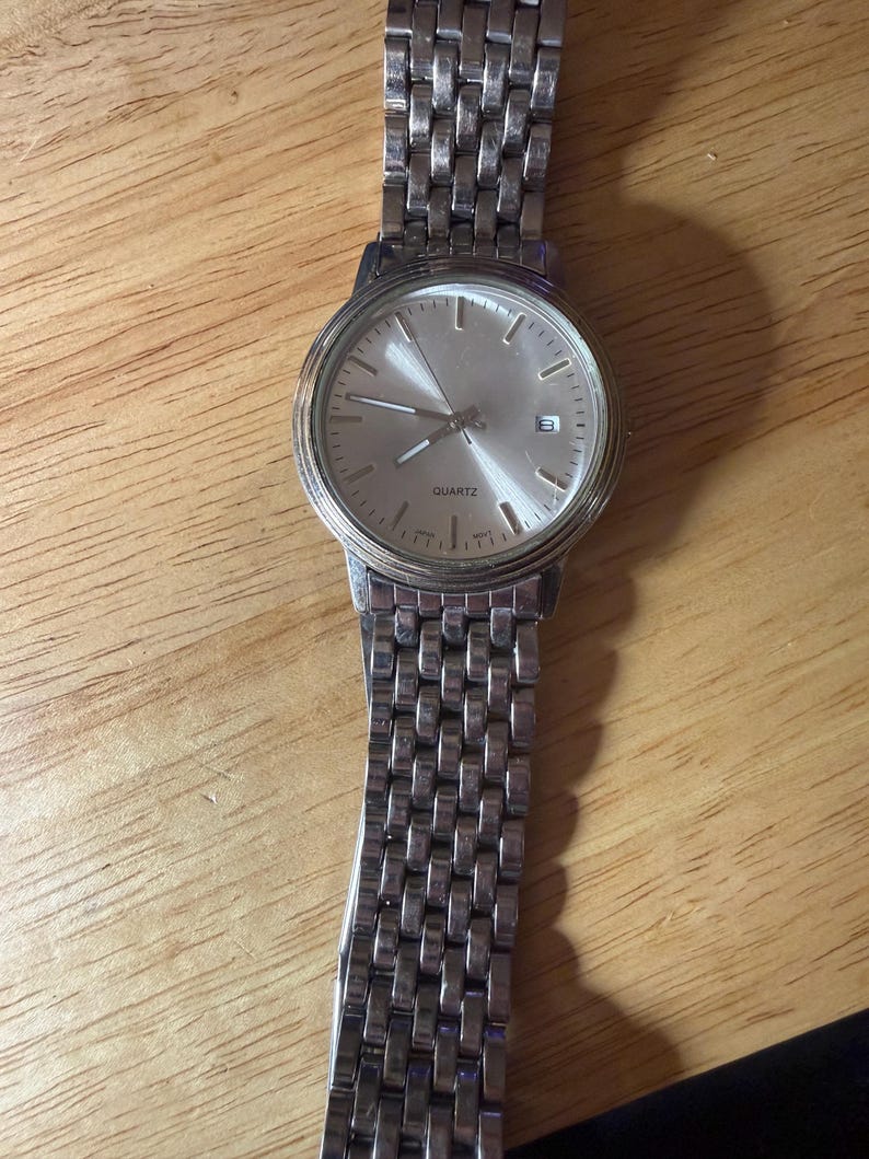 May include: Silver-toned quartz watch with a rectangular link band. The watch face is silver with black hour markers and hands. The word "QUARTZ" is printed on the watch face. The date is displayed at the 3 o'clock position.