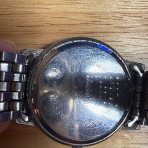 May include: Close-up of a stainless steel watch with a silver band. The watch back is visible, with text that reads "Stainless Steel Back" and "Japan Movement Base". The watch is on a wooden surface.