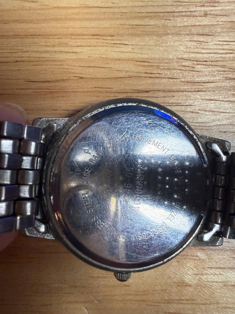 May include: Close-up of a stainless steel watch with a silver band. The watch back is visible, with text that reads "Stainless Steel Back" and "Japan Movement Base". The watch is on a wooden surface.
