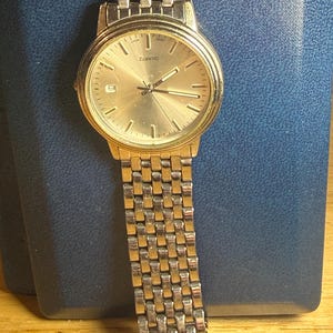 May include: A gold-toned quartz watch with a rectangular link band. The watch face is gold with black hands and markers. The watch has a date display at the 6 o'clock position. The watch is displayed on a blue surface.