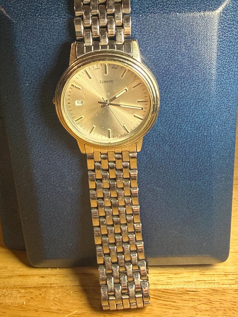 May include: A gold-toned quartz watch with a rectangular link band. The watch face is gold with black hands and markers. The watch has a date display at the 6 o'clock position. The watch is displayed on a blue surface.