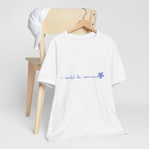 Puede incluir: Camiseta blanca con la frase "i could be meaner" en escritura azul claro y un gráfico de flor azul. La camiseta está colgada en una percha de madera, con una gorra de béisbol blanca sobre una silla de madera.