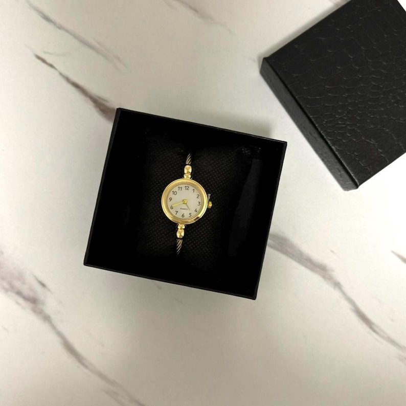 K&ouml;nnte beinhalten: Eine goldfarbene Ringuhr mit wei&szlig;em Zifferblatt und schwarzen Ziffern, pr&auml;sentiert in einer schwarzen Schachtel. Die Uhr hat ein filigranes Design mit einem d&uuml;nnen Band. Das Zifferblatt ist rund und hat goldene Zeiger.