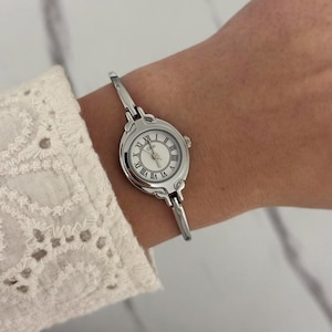 May include: A silver wristwatch with a white face and Roman numeral hour markers. The watch has a slender, hinged band and is worn on a wrist. The watch face has the word "Gedi" printed on it.