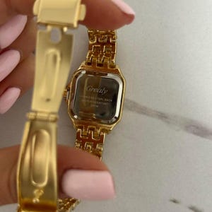 Puede incluir: Un reloj de pulsera dorado con una esfera rectangular y una correa de eslabones a juego. La esfera del reloj tiene el nombre de la marca "Grealy" impreso, junto con el texto "STAINLESS STEEL BACK WATER RESISTANT 3ATM".