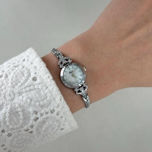 May include: A silver-toned wristwatch with a white face and decorative crystal accents. The watch features a delicate, ornate band and a small, round face. The watch is worn on a wrist, showcasing its elegant design.