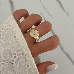 May include: A gold-colored watch ring with a cream-colored face and a small, silver-colored hand. The ring is designed to look like a wristwatch and is worn on a finger.