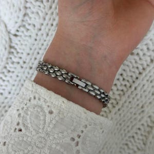 May include: A silver-toned bracelet with an oval link design is fastened on a wrist. The bracelet is made of metal and has a secure clasp. The wrist is partially visible, with a white, textured fabric in the background.