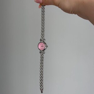 May include: A silver-toned wristwatch with a pink face and a linked band. The watch has a round face with small hour markers and a delicate, chain-style band. The watch is held up against a plain background.