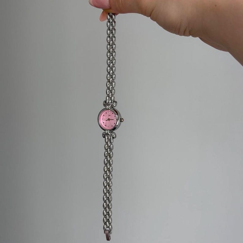May include: A silver-toned wristwatch with a pink face and a linked band. The watch has a round face with small hour markers and a delicate, chain-style band. The watch is held up against a plain background.