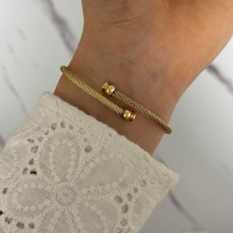 K&ouml;nnte beinhalten: Ein goldfarbenes, offenes Armband. Das Armband hat ein gedrehtes Seildesign mit abgerundeten Goldakzenten an jedem Ende. Das Armband wird an einem Handgelenk gezeigt, mit einem wei&szlig;en Spitzensaum im Hintergrund.