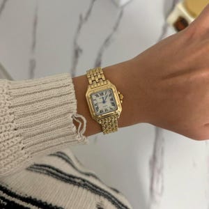 Puede incluir: Un reloj de pulsera dorado con una esfera cuadrada y una correa dorada a juego. La esfera del reloj es blanca con n&uacute;meros romanos negros y agujas doradas. El reloj se lleva en la mu&ntilde;eca.