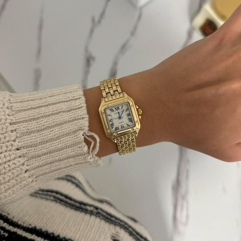 Puede incluir: Un reloj de pulsera dorado con una esfera cuadrada y una correa dorada a juego. La esfera del reloj es blanca con n&uacute;meros romanos negros y agujas doradas. El reloj se lleva en la mu&ntilde;eca.