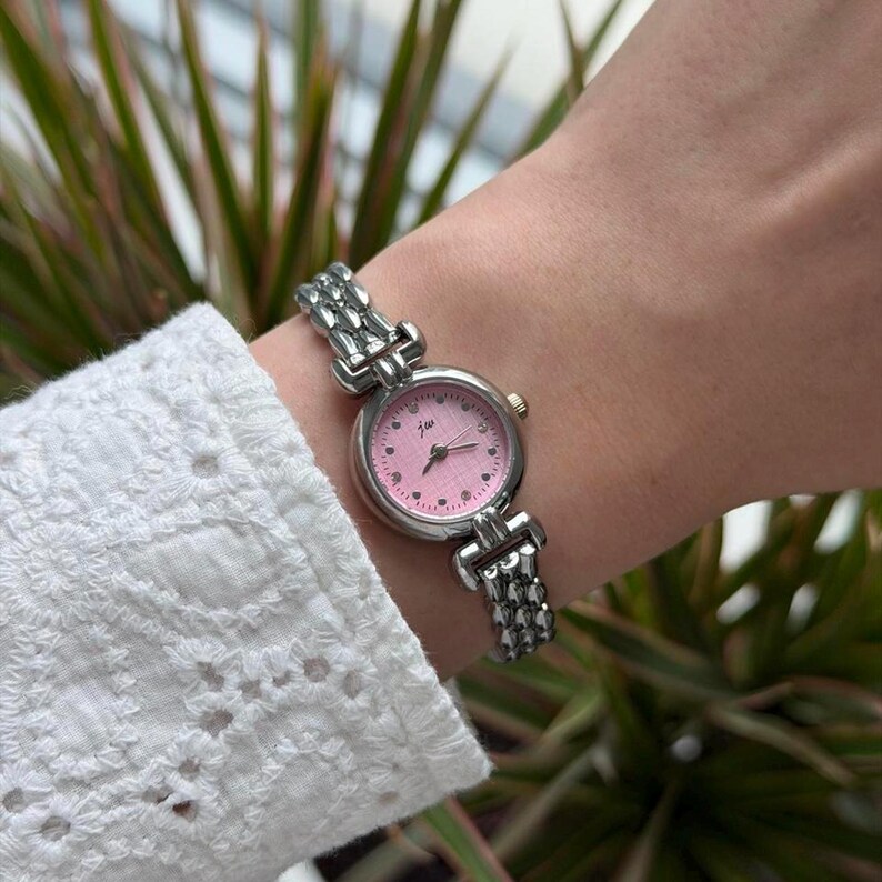 May include: A silver-toned wristwatch with a pink face and a linked band is displayed. The watch face features small heart-shaped markers and the initials "jw". The watch is worn on a wrist, with a white embroidered sleeve in the background.