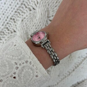 May include: A silver wristwatch with a pink face and a silver band. The watch has a round face with small crystals around the edge. The band is made of small, connected silver links.