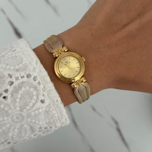 Könnte beinhalten: Eine goldfarbene analoge Armbanduhr mit rundem Zifferblatt, einem einzelnen Stein-Index auf der Zwölf-Uhr-Position, schmalen Zeigern und einem strukturierten Mesh-Armband. Das Zifferblatt zeigt die Buchstaben BS und den Text Bee Sister.