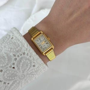 May include: A gold-toned rectangular wristwatch with a cream-colored face and Roman numeral hour markers. The watch has a gold band with a ribbed design. The watch is on a wrist, with a white lace sleeve in the background.