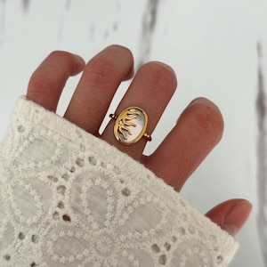 May include: Gold-toned ring featuring an oval-shaped mother-of-pearl center with a sun design. A small diamond accent is set near the sun. The ring is displayed on a finger, showcasing its design and details.