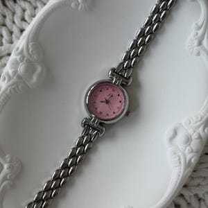 May include: A silver-toned wristwatch with a pink face and a linked band. The watch face has small hour markers and delicate hands. The watch is displayed on a white, decorative tray.