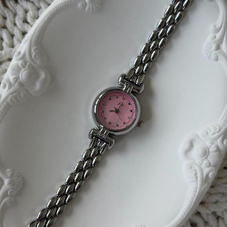 May include: A silver-toned wristwatch with a pink face and a linked band. The watch face has small hour markers and delicate hands. The watch is displayed on a white, decorative tray.