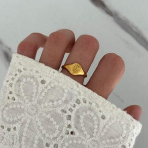 May include: A gold-colored signet ring with a floral design. The ring is oval-shaped and features a detailed engraving of a small bouquet. The ring is worn on a finger, with a white lace sleeve in the background.