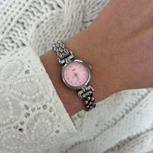 May include: A silver-toned wristwatch with a pink face is displayed on a wrist. The watch has a round face with small dots marking the hours and delicate hands. The band is made of connected, rounded metal links.