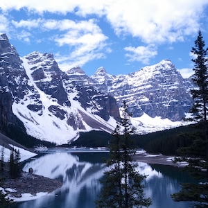 Puede incluir: Un paisaje escénico con montañas cubiertas de nieve reflejadas en un lago tranquilo. El cielo es de un azul brillante con nubes blancas dispersas. Los árboles de hoja perenne enmarcan el lago y las montañas, creando una escena natural serena.