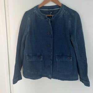 May include: A blue jacket with a textured, waffle-like pattern. The jacket has a round collar, button closures, and two front pockets. It is hanging on a wooden hanger.