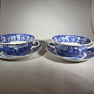 Könnte beinhalten: Zwei blau-weiße Porzellan-Suppenschalen mit passenden Untertassen. Die Schalen haben zwei Griffe und ein Blumen- und Landschaftsdesign. Die Untertassen weisen ein passendes Landschaftsdesign auf. Das Set wird auf einer weißen Oberfläche präsentiert.